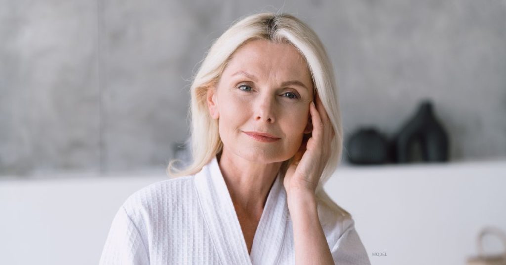 mature beautiful woman in white robe, touching her face, admiring her post facelift results (MODEL)